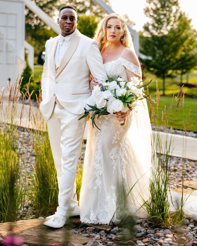 A bride and groom in a cream and champagne tuxedo standing outside near a garden.