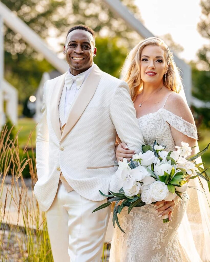 A bride and groom in a cream and champagne tuxedo standing outside near a garden.