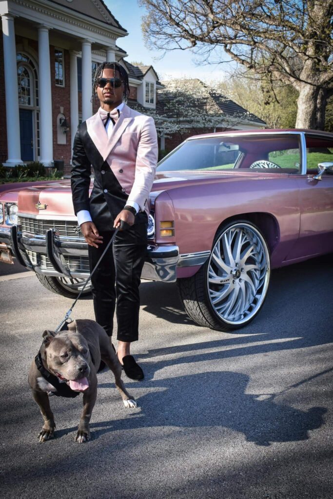 A man wearing a pink and black suit standing in front of a car with a dog on a leash.