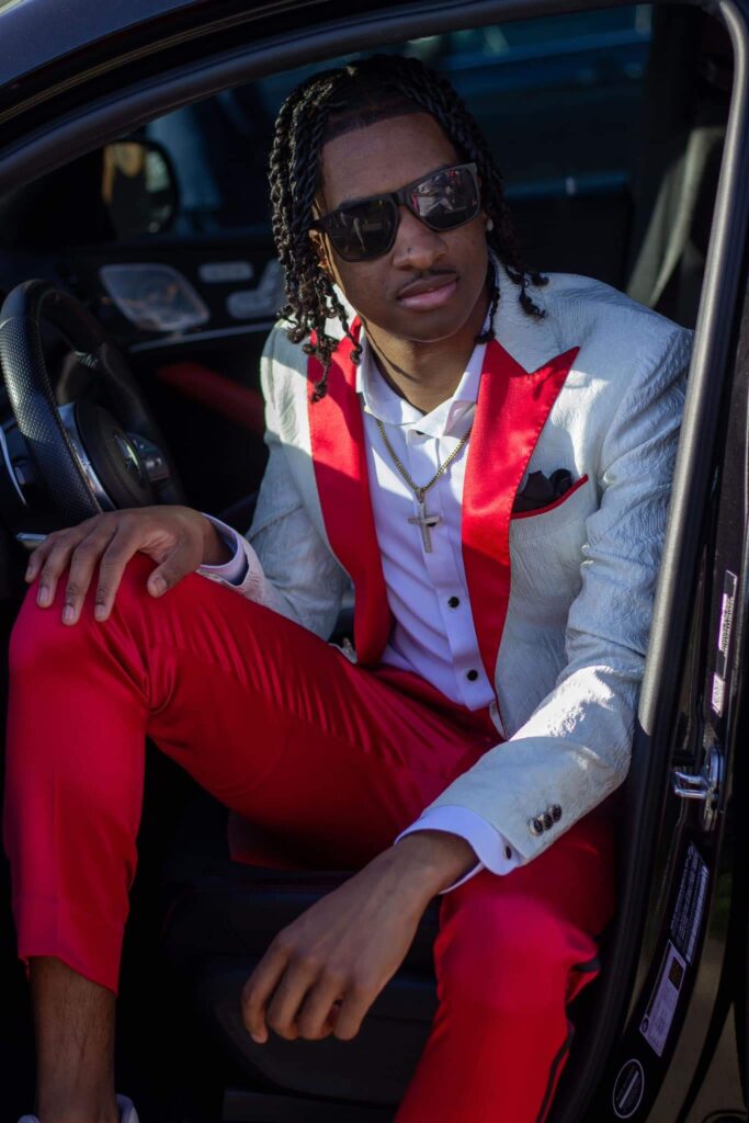 A boy sitting in a car wearing a cream and red tuxedo sitting in a car