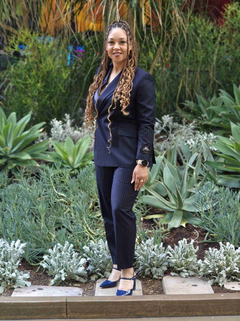 DeNira Dedeaux in a custom blue suit standing in front of flowers. Custom Suits Kansas City